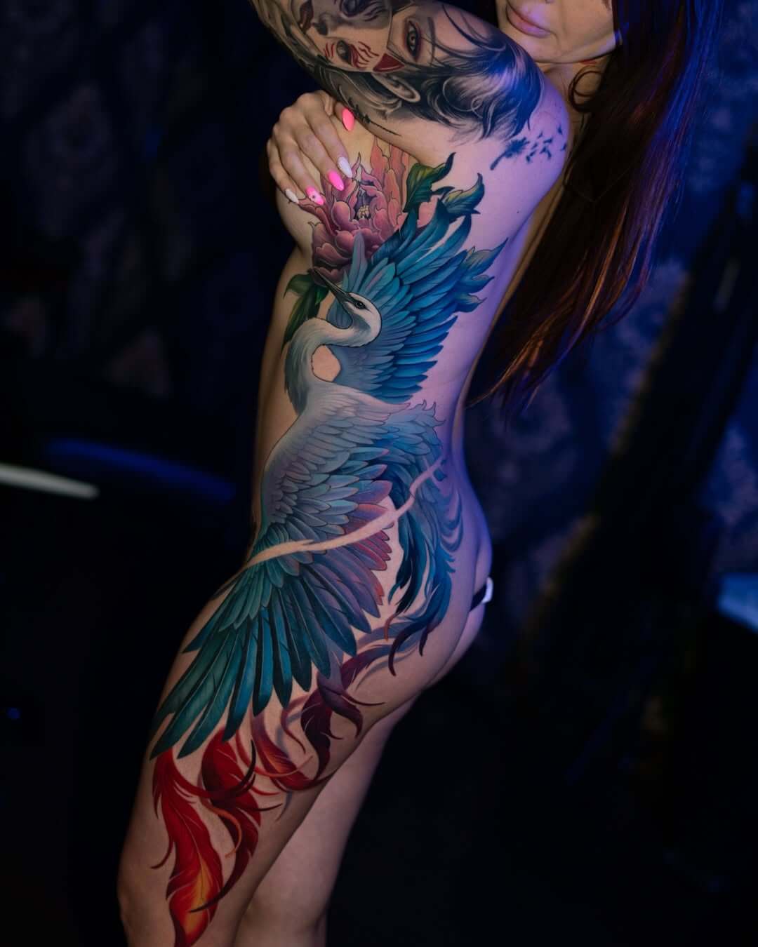 Vibrant blue crane and pink flower tattoo on female ribs and hip. High-detail Neo-traditional bird tattoo with smooth color gradients and botanical elements.