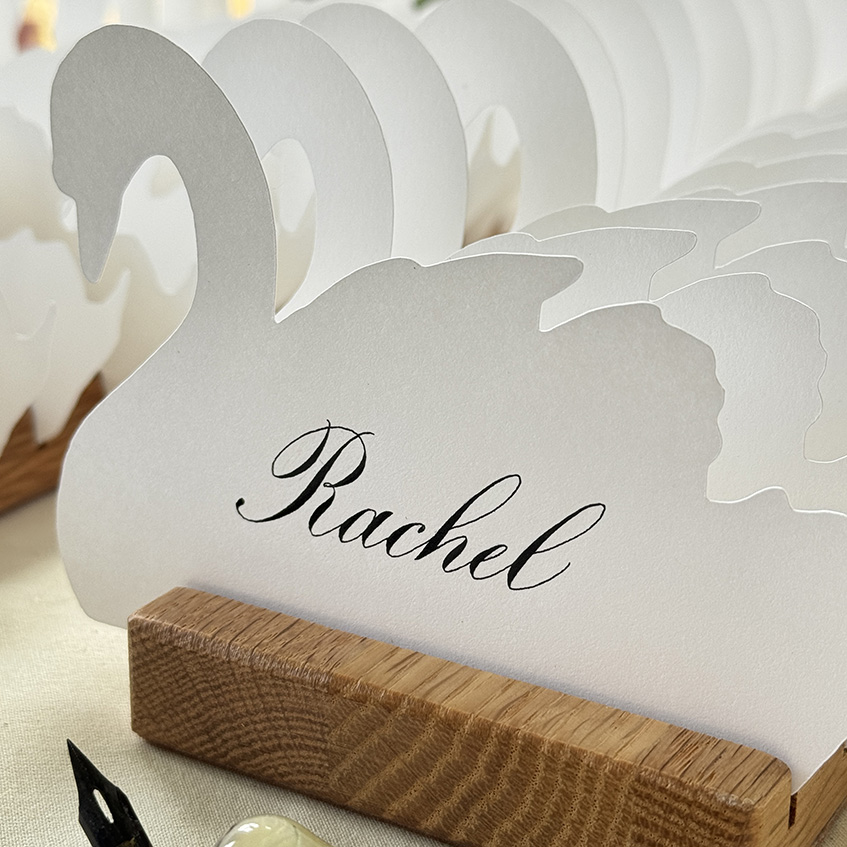 Swan-shaped wedding place card with black Copperplate calligraphy in a wooden holder.