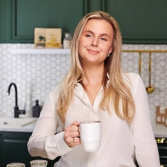 Портретная фотография улыбающейся молодой женщины со светлыми прямыми волосами, в белой рубашке, смотрящей прямо в камеру и держащей белую керамическую кружку в правой руке. На заднем плане видна часть кухни с тёмно-зелёными шкафами, белой столешницей и металлическими аксессуарами.