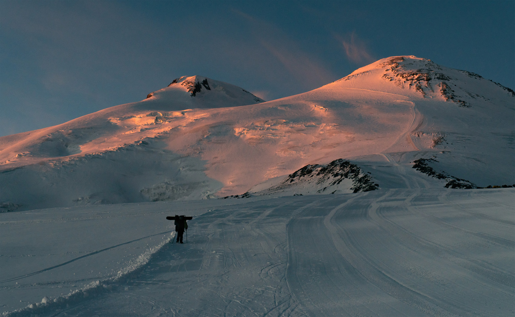 Фотография южного склона горы Эльбрус освещенные красными лучами закатного солнца. Сноубордист несет снаряжение по склону Эльбруса. Фотограф Севальнев Ян