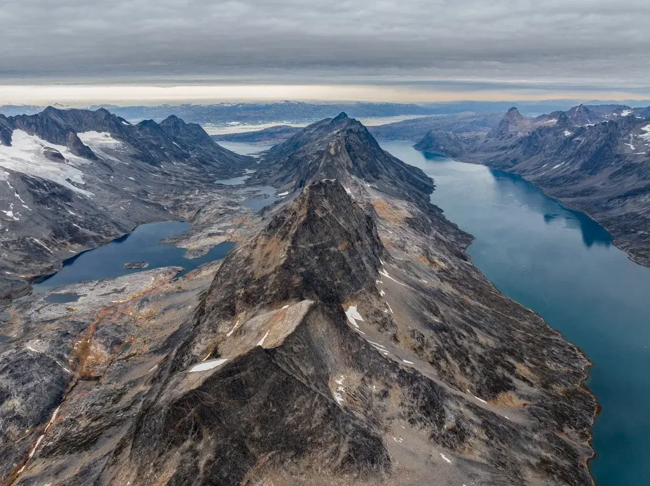 Aerial view of Greenlands dramatic mountain range and deep fjords with rugged peaks glacial lakes and dark Arctic waters stretching beneath a cloudy sky