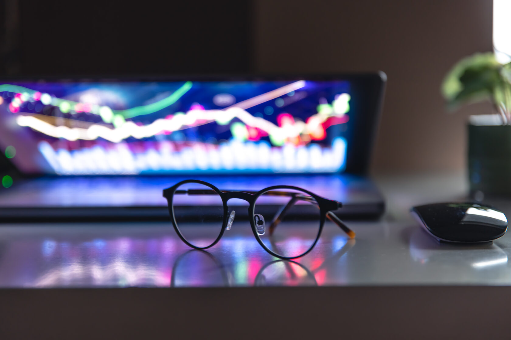 gafas sobre la mesa y al fondo gráficos de trading en la pantalla de una laptop
