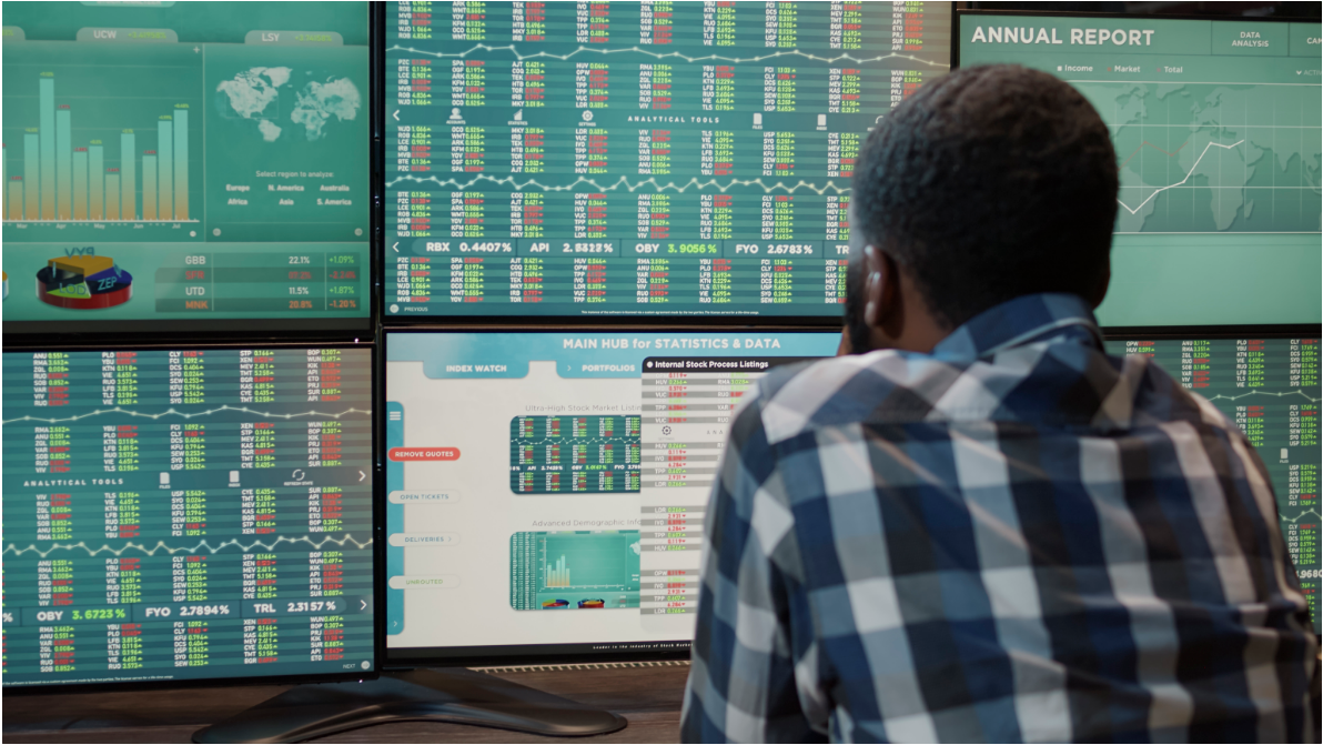 A man viewed from behind, looking at six monitors displaying stock charts and trading signals