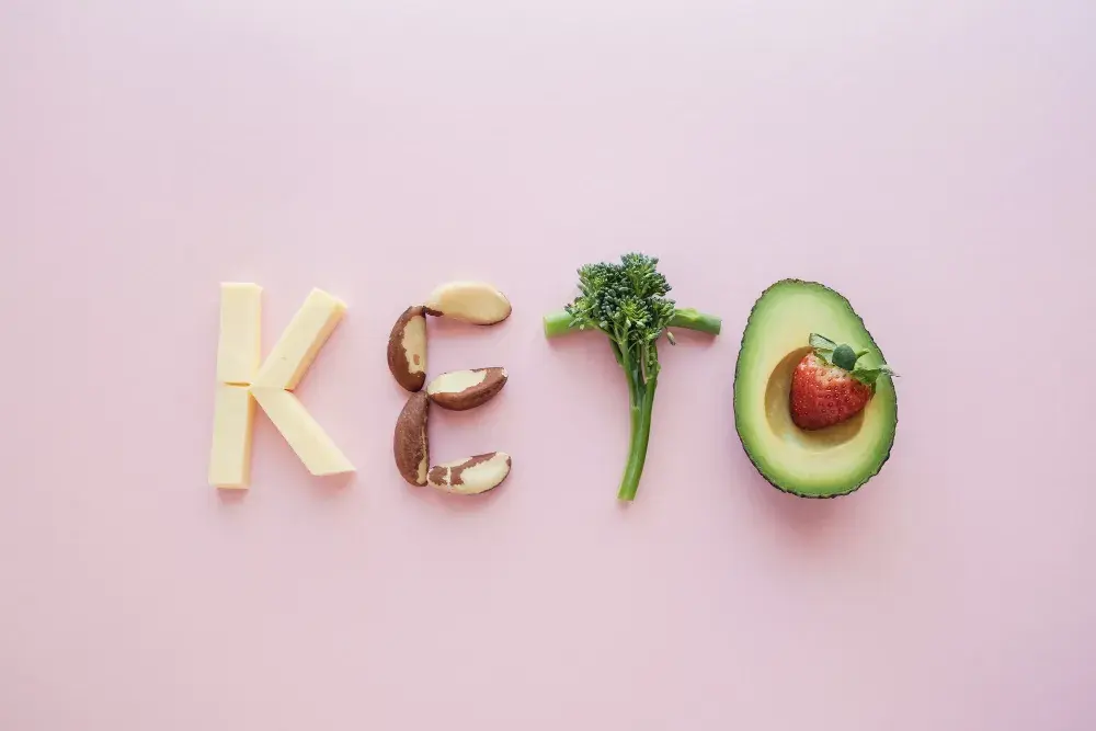 Написано слово “KETO” из продуктов: сыр, орехи, брокколи, авокадо и клубника на розовом фоне.