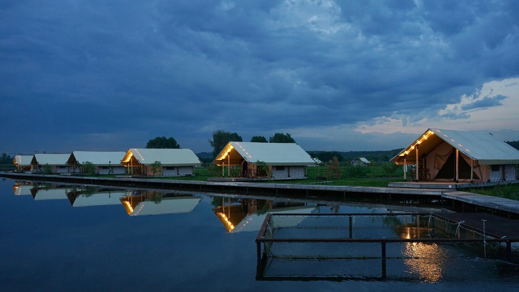 Safari tents lit up after dark, reflected in the water at Ostrov Glamping