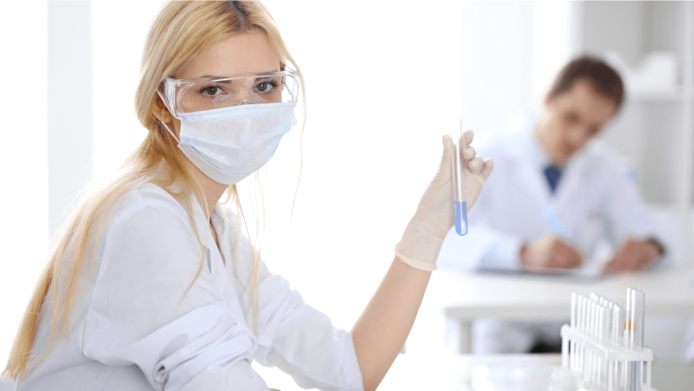 Женщина-ученый в защитной маске и очках держит перед собой пробирку с голубой жидкостью