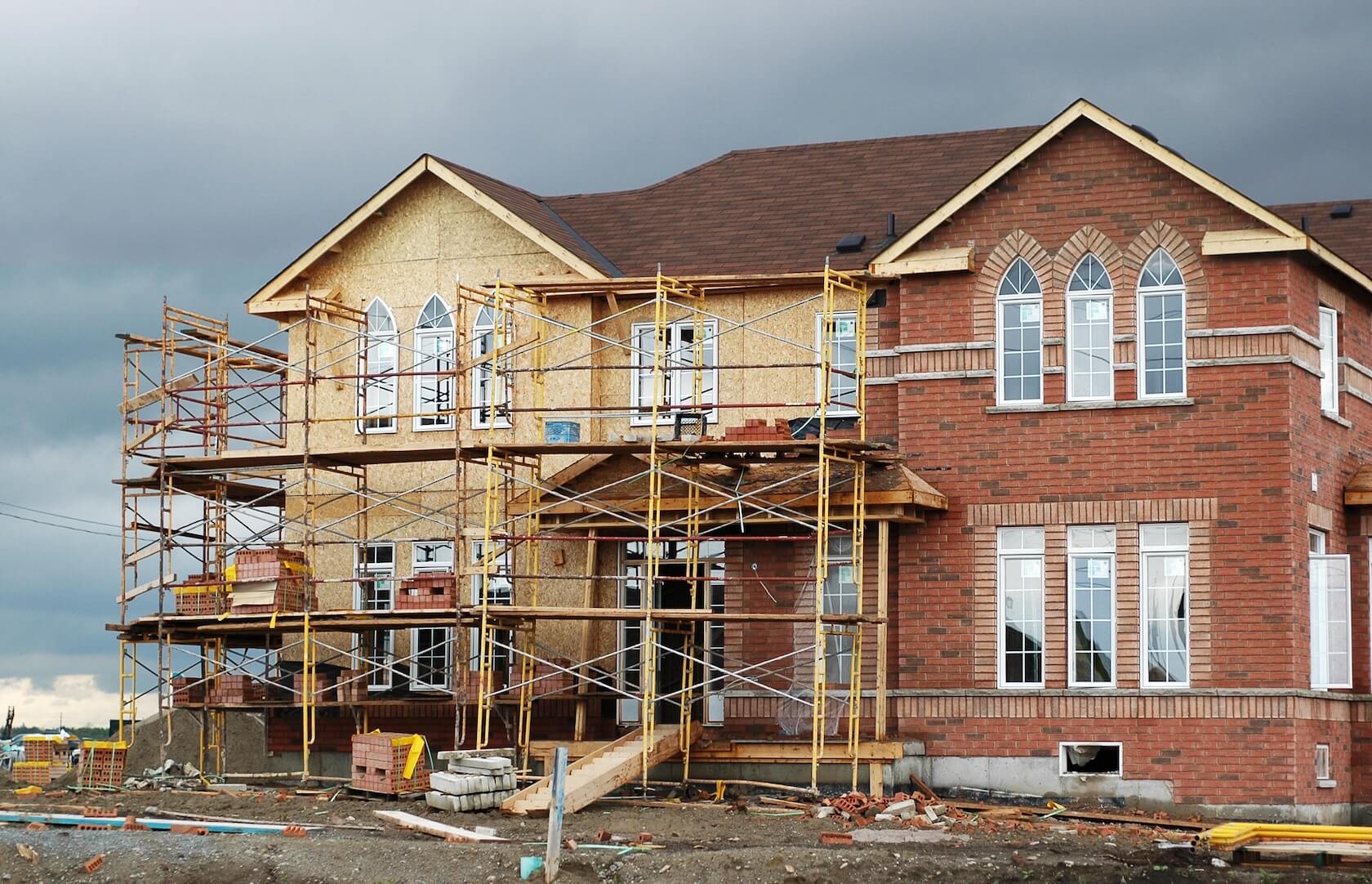 Частный дом в процессе облицовки фасада. На одной части стены уже уложен облицовочный кирпич, другая покрыта плитами ОСП и подготовлена к работам. Вокруг установлены строительные леса и материалы.