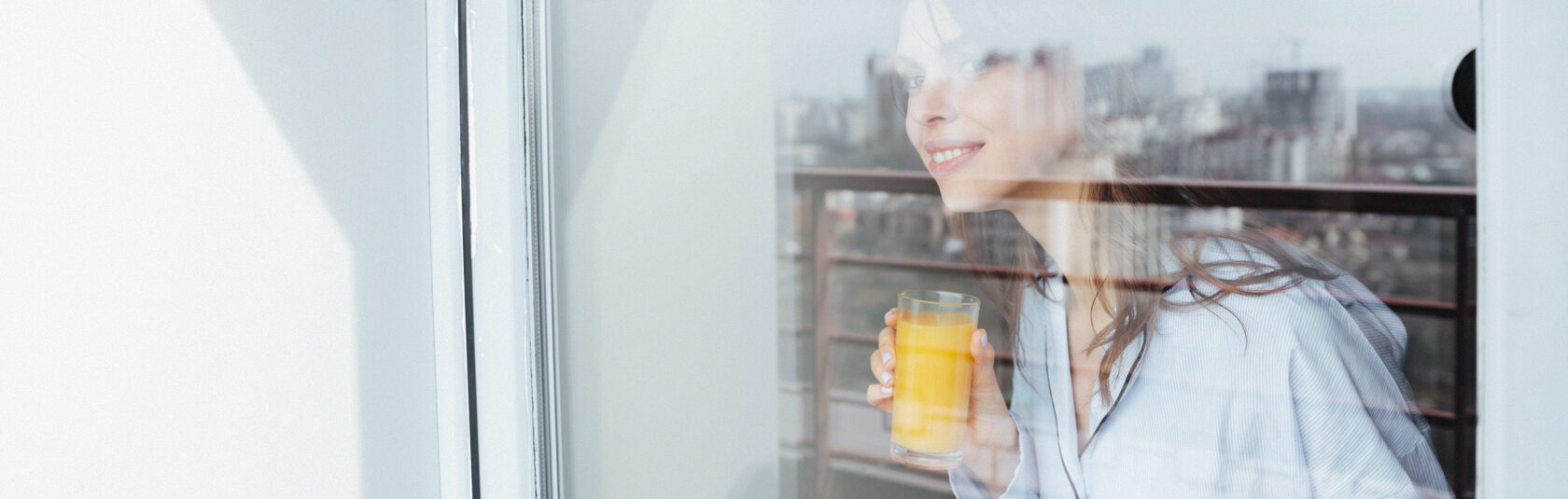 Девушка стоит у окна с чашкой напитка и смотрит на город через чистое стекло