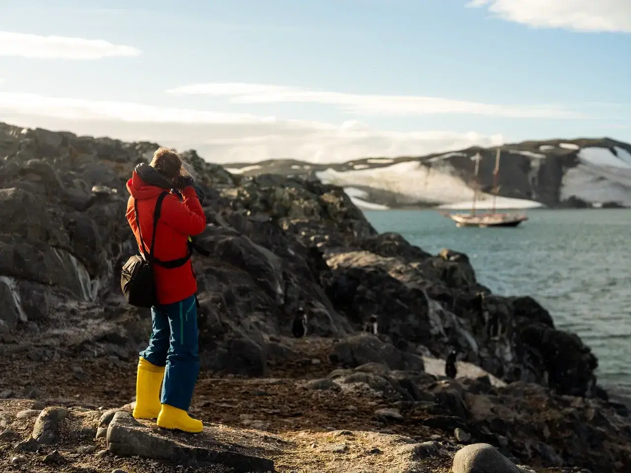A photographer captures Antarcticas breathtaking landscapes during a MoreTime Expeditions sailing expedition. Standing on a rocky shoreline surrounded by penguins with a traditional schooner anchored in the bay and snow-covered mountains in the background