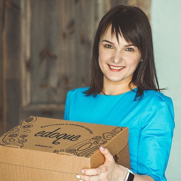 Портретная фотография улыбающейся молодой женщины с темными волосами, собранными в короткую стрижку с челкой, в синем платье, держащей открытую картонную коробку с надписью 'edoque' и иллюстрациями фруктов и овощей. На заднем плане видна размытая деревянная дверь.