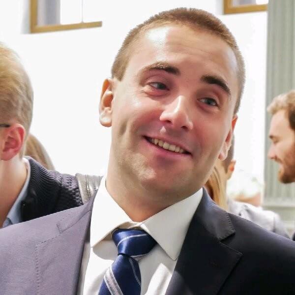 Портретная фотография улыбающегося молодого мужчины со светлыми короткими волосами, в синем деловом костюме, белой рубашке и темно-синем галстуке в светлую полоску. Мужчина смотрит в левый верхний угол, демонстрируя открытую улыбку, на заднем плане видны размытые силуэты других людей.