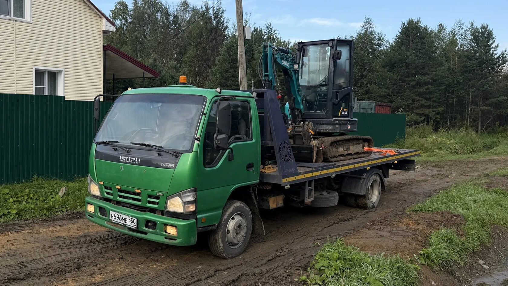 Эвакуатор Isuzu перевозит мини-экскаватор через дачный посёлок