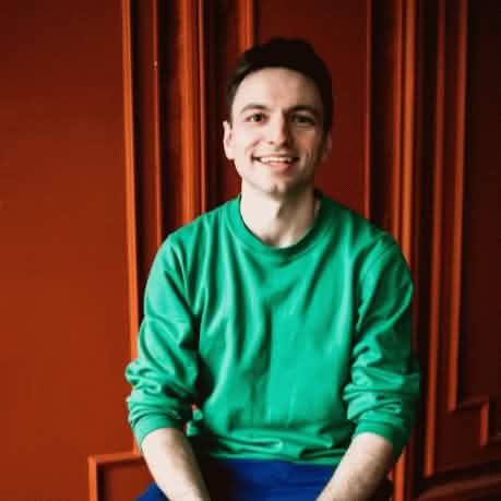 Портретная фотография улыбающегося мужчины с темными волосами, чисто выбритый, в ярко-зеленом свитере, сидящего на фоне вертикальной красной стены.