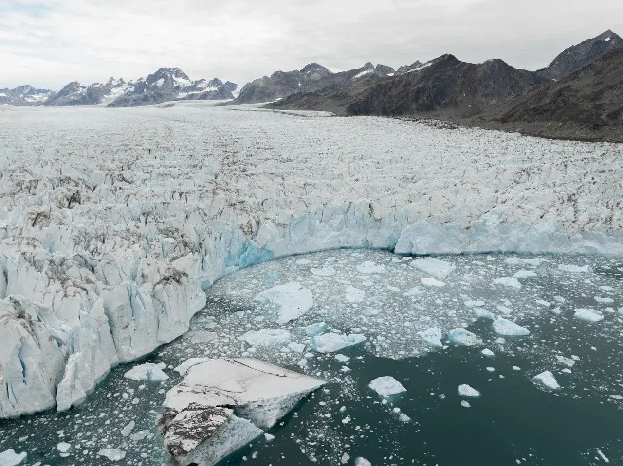Aerial view of a massive Greenland glacier flowing into an icy lagoon filled with floating ice chunks, surrounded by rugged mountain peaks and the dramatic Arctic landscape.