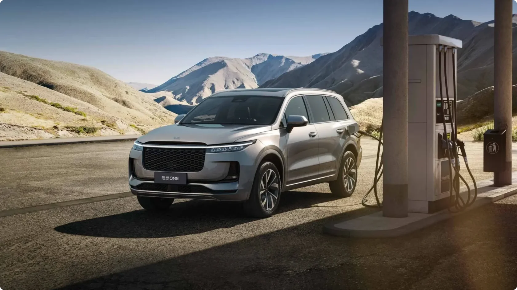 Гибридный кроссовер Li Auto ONE серебристого цвета на зарядке у колонки на фоне горного пейзажа, процесс зарядки батареи.