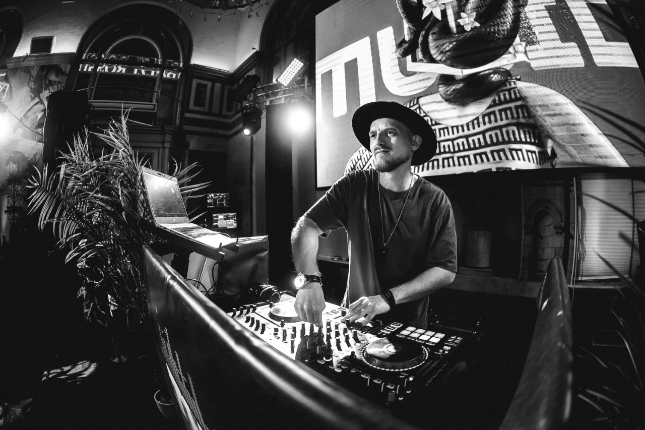 Чёрно-белая фотография DJ ROZHKOV в шляпе за пультом на оформленной сценической площадке.