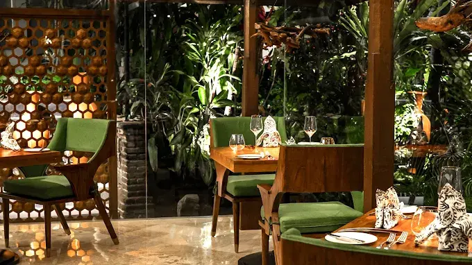 An upscale top dinner in Ubud set in a lush indoor-outdoor garden space, detailed with elegant wooden tables, chic green chairs, and a highly romantic evening ambiance.
