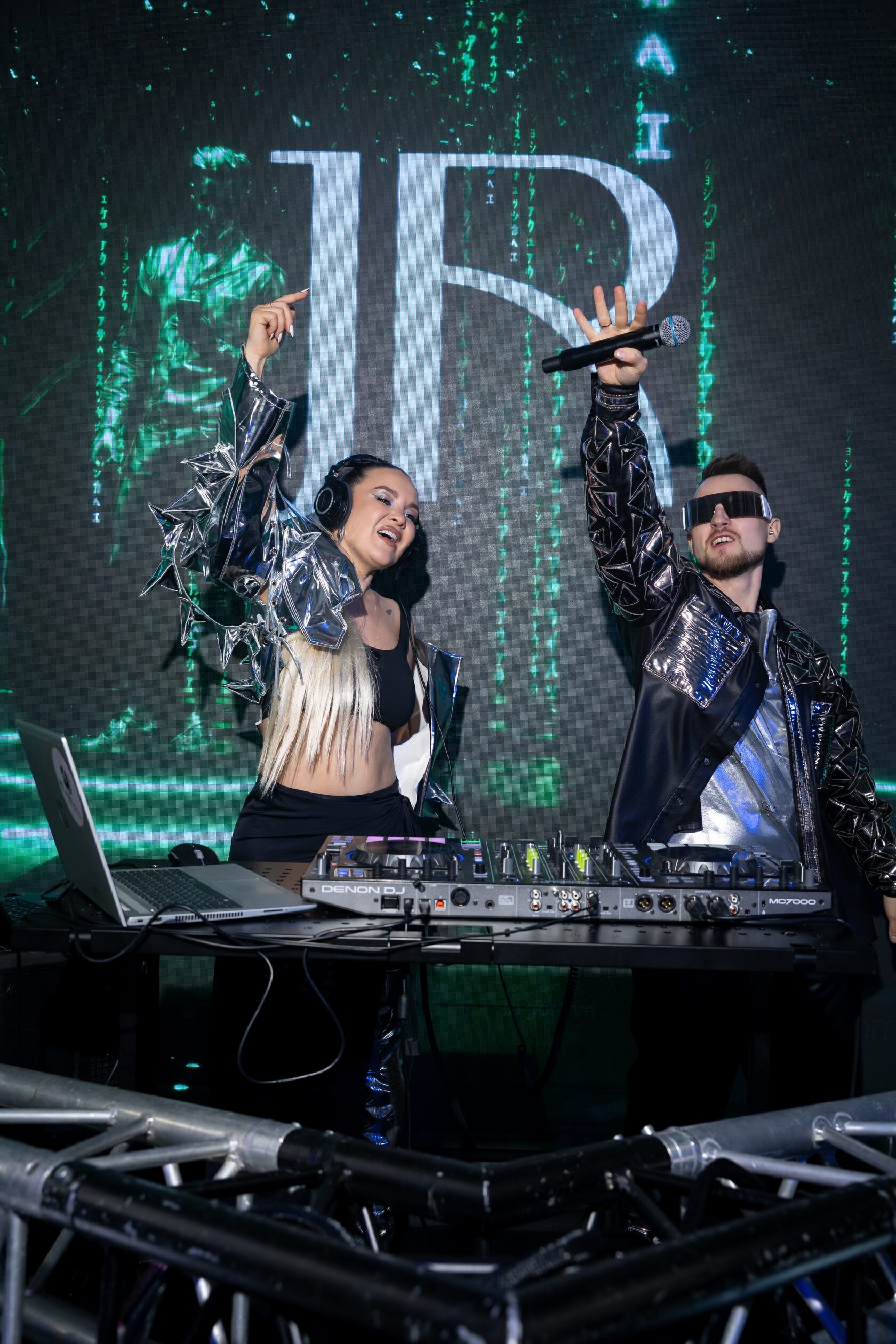 DJ ROZHKOV и вокалистка в серебристом сценическом образе поднимают руки на сцене рядом с пультом.