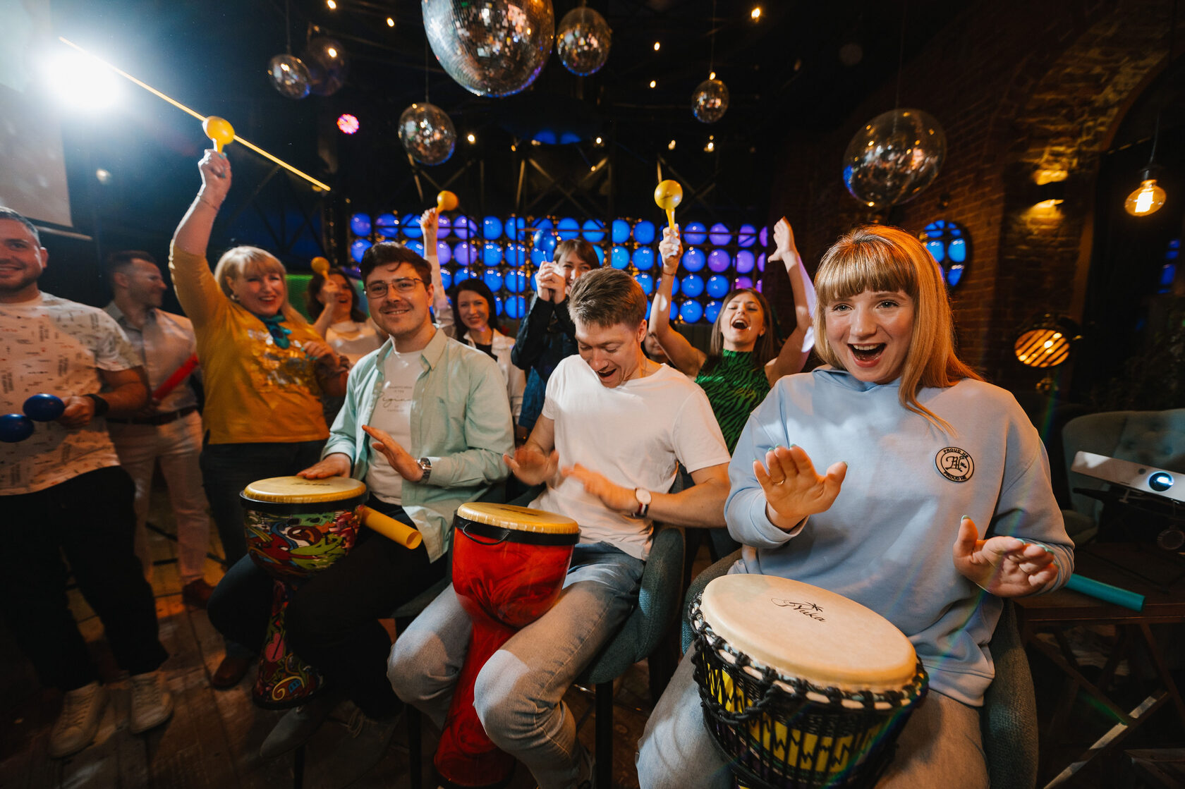 Фотографии с вечеринки: мужчины и девушки играют на африканских баранах под задорный ритм ведущего. На заднем плане девушки аккомпанируют на маракасах. Барабанное шоу — лучшее, что можно заказать на запоминающееся мероприятие