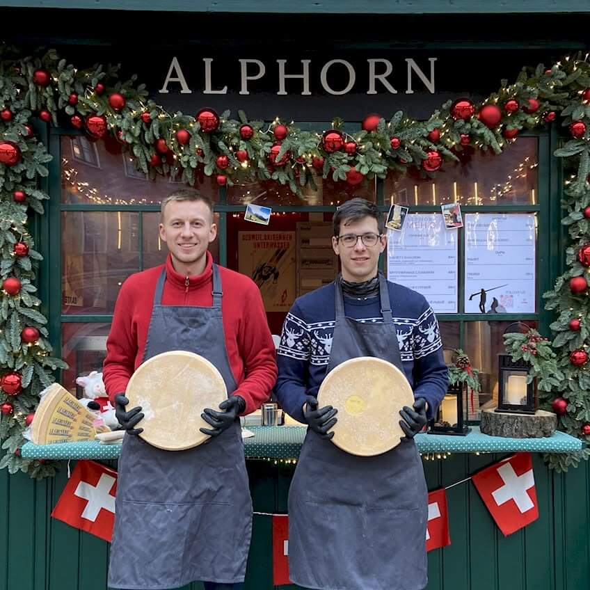 Фотография двух молодых мужчин, стоящих перед витриной магазина 'ALPHORN', украшенной новогодним декором. Мужчина слева в красной толстовке и сером фартуке, а мужчина справа в синем свитере с белыми узорами и сером фартуке. Оба мужчины держат круглые головки сыра и одеты в черные перчатки. На заднем фоне видны флаги Швейцарии.