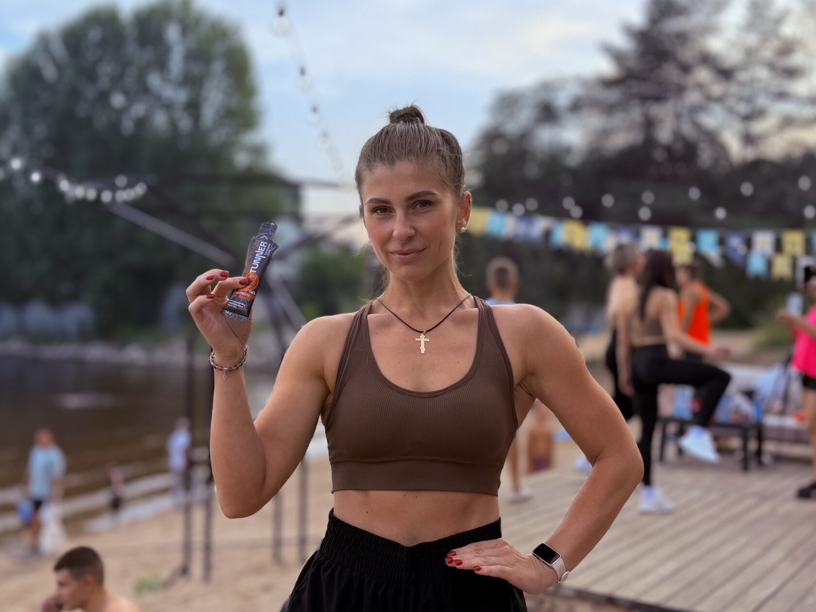 Девушка в спортивной форме держит в руке упаковку высокоуглеводного геля TUNNER на фоне участников фитнес-фестиваля