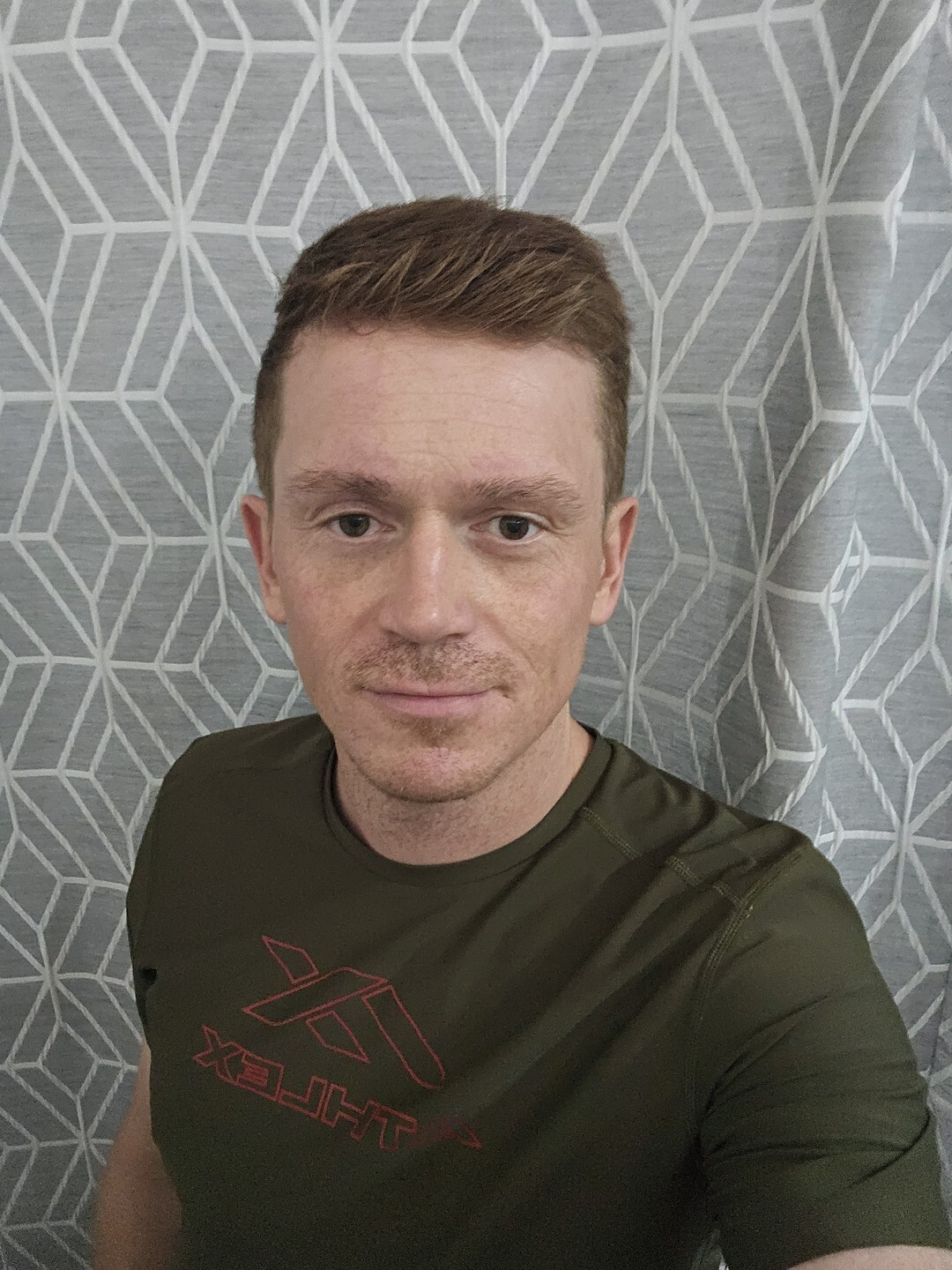 Павел Ростовцев, крупный портрет у серой стены с узором, оливковая футболка, лёгкая улыбка