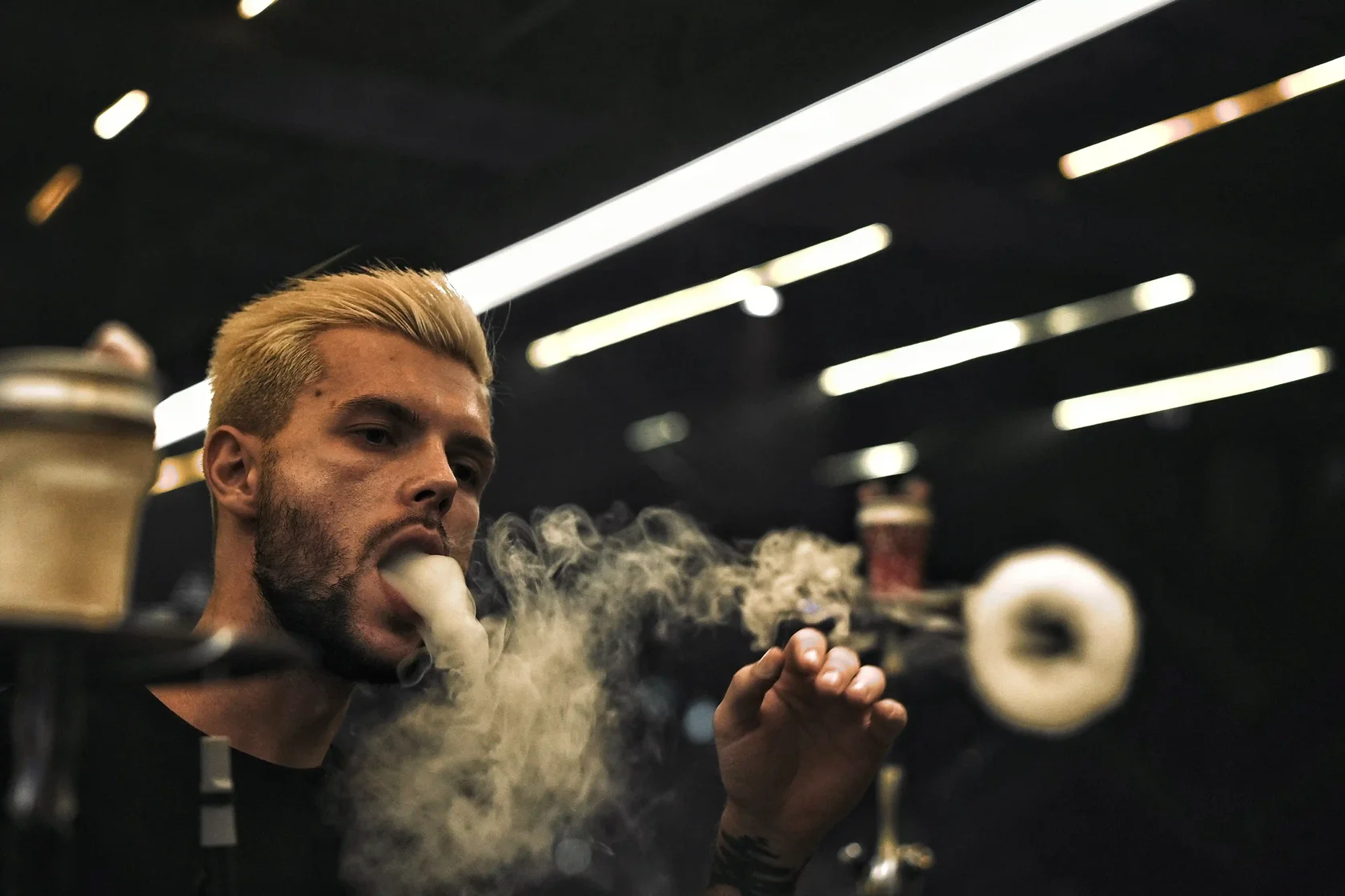 A close-up of a stylish man exhaling a perfect thick ring of shisha smoke in a modern dimly lit lounge setting.