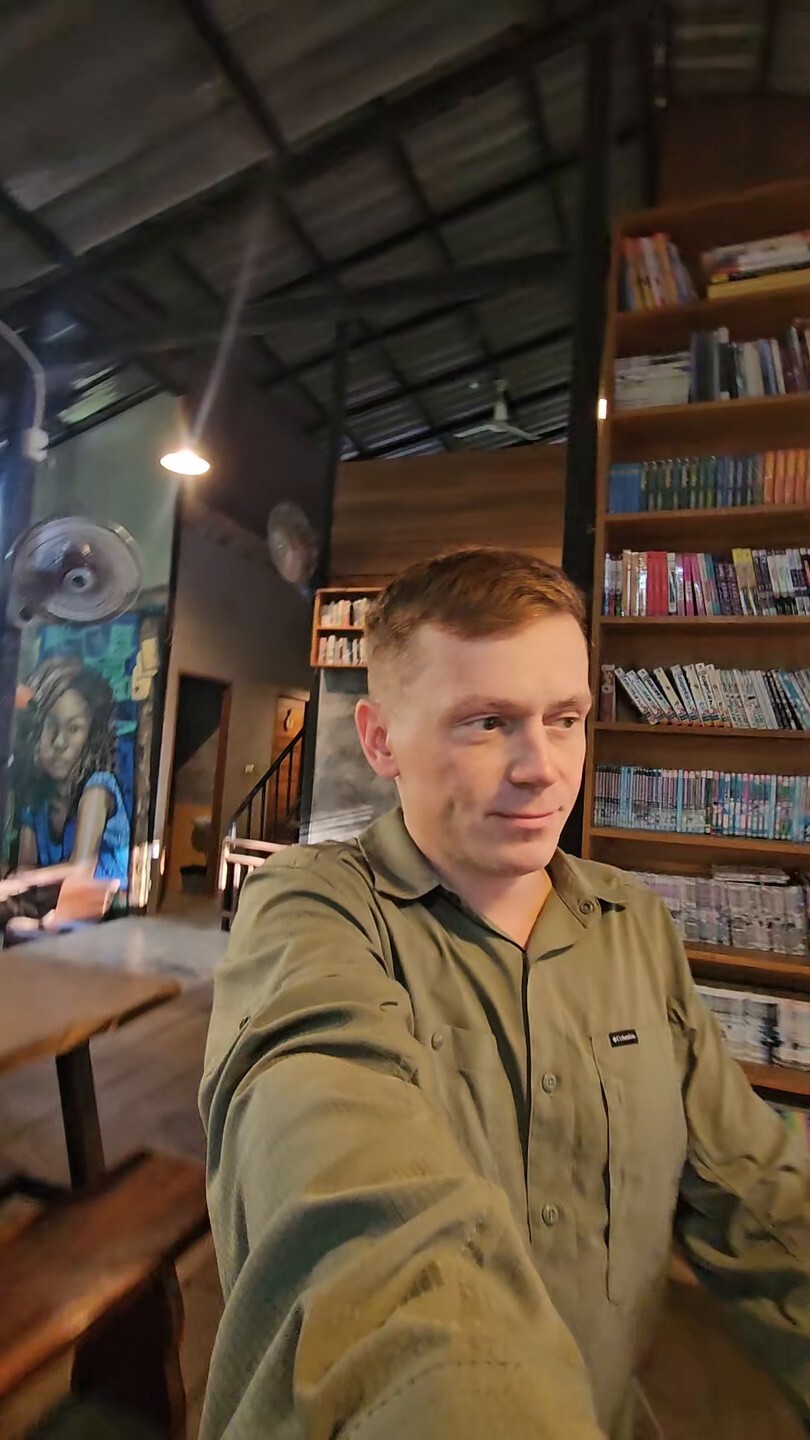 Павел Ростовцев делает селфи в кафе с книжными стеллажами, рубашка хаки