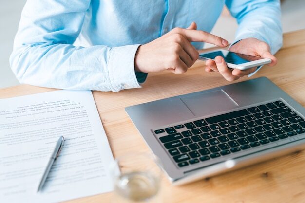 Young employee in blue shirt sitting by wooden table in front of laptop and scrolling in smartphone during communication