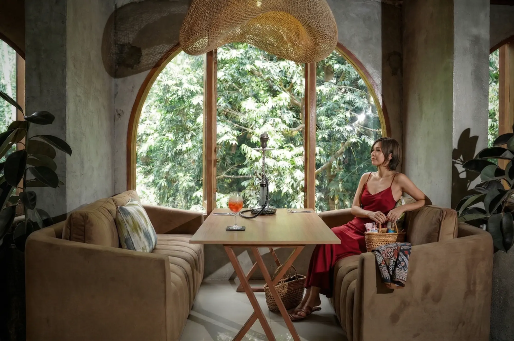 A stylish woman in a red dress relaxing with a drink and shisha by a beautiful arched window, winding down after a busy day exploring the Ubud Art Market.