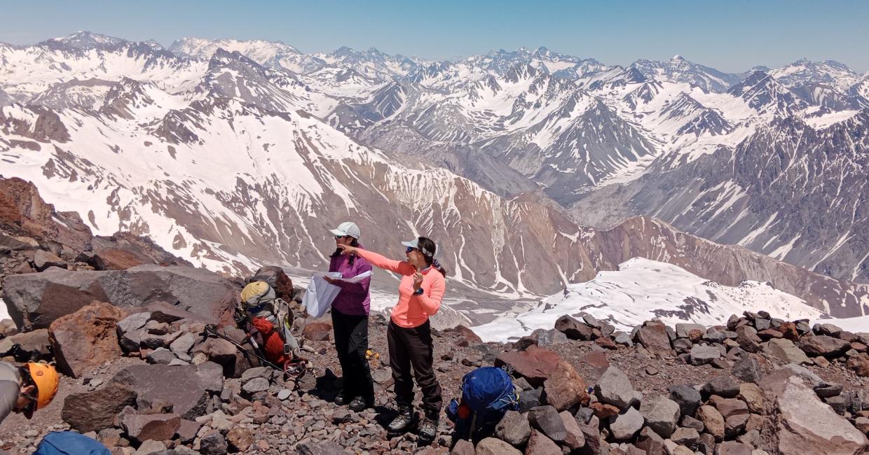 Group of mountaineers on rocky high mountain terrain, with snowy peaks in the background, assessing the route and pointing toward the landscape during a guided activity.