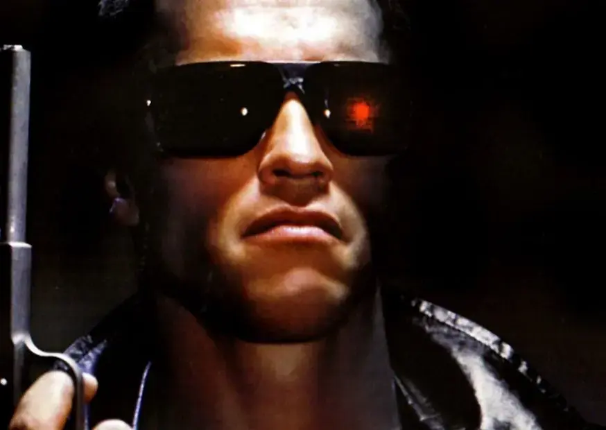 Кадр из фильма «Терминатор»: мужчина в чёрной куртке и солнцезащитных очках держит оружие