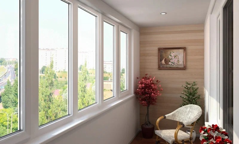 Застеклённый балкон с белыми пластиковыми окнами и видом на город летом, внутри — уютное кресло, растения и картина на стене.