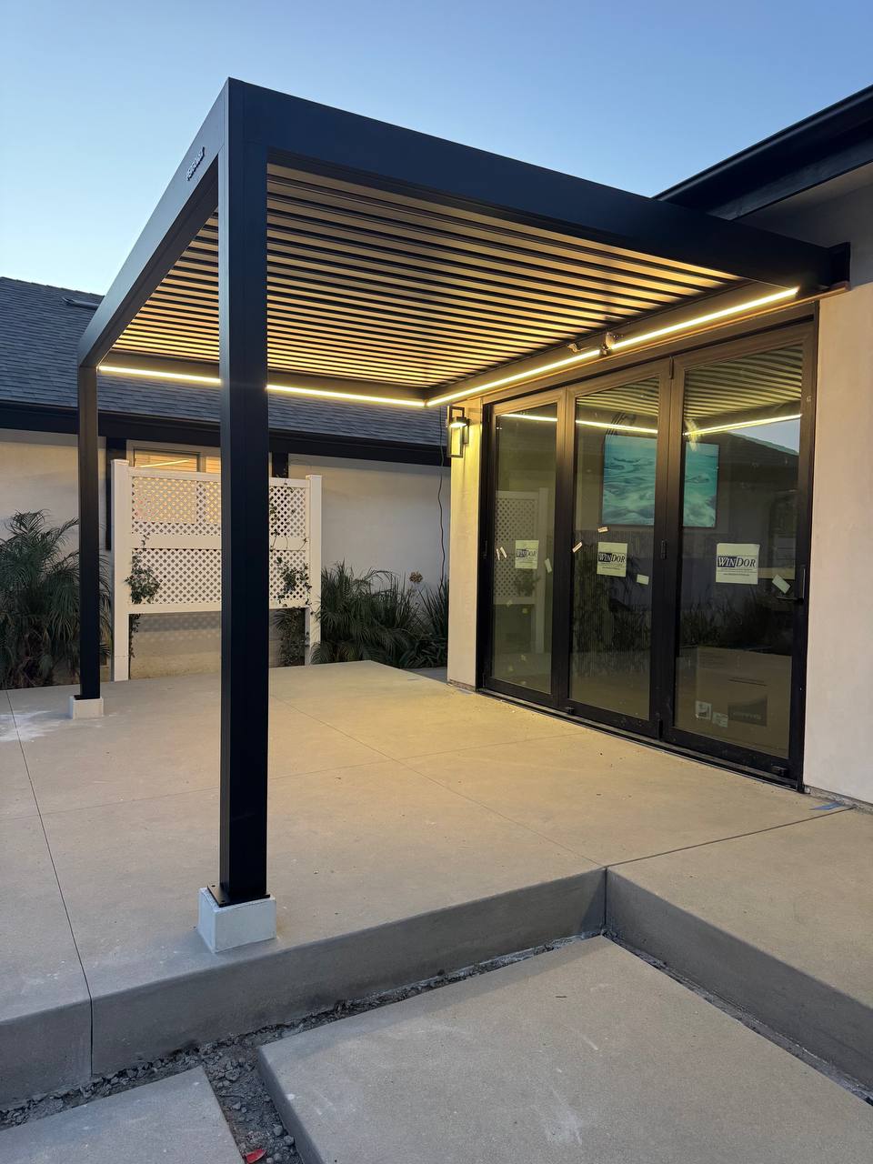 Black attached pergola with perimeter LEDs switched on against a blue evening sky.