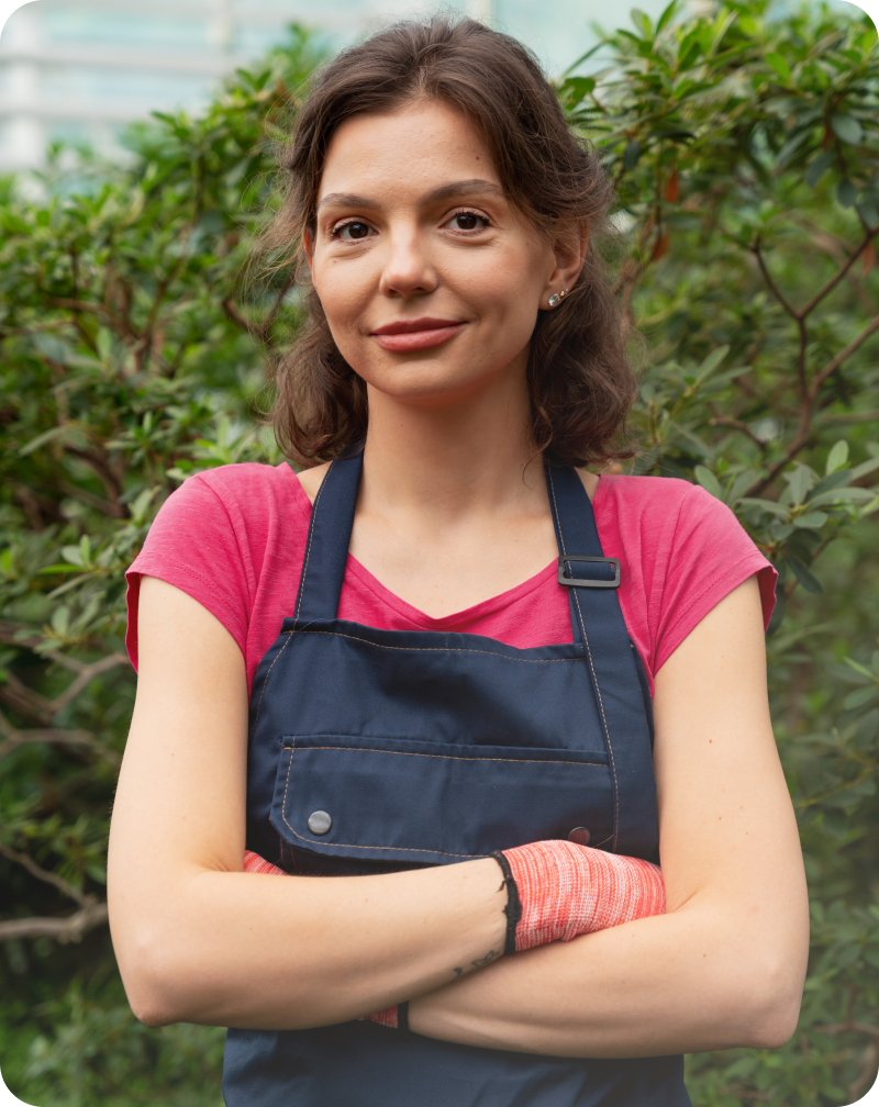 Женщина в синем фартуке стоит перед камерой на фоне зеленой растительности. Она одета в розовую футболку и держит руки скрещенными на груди.