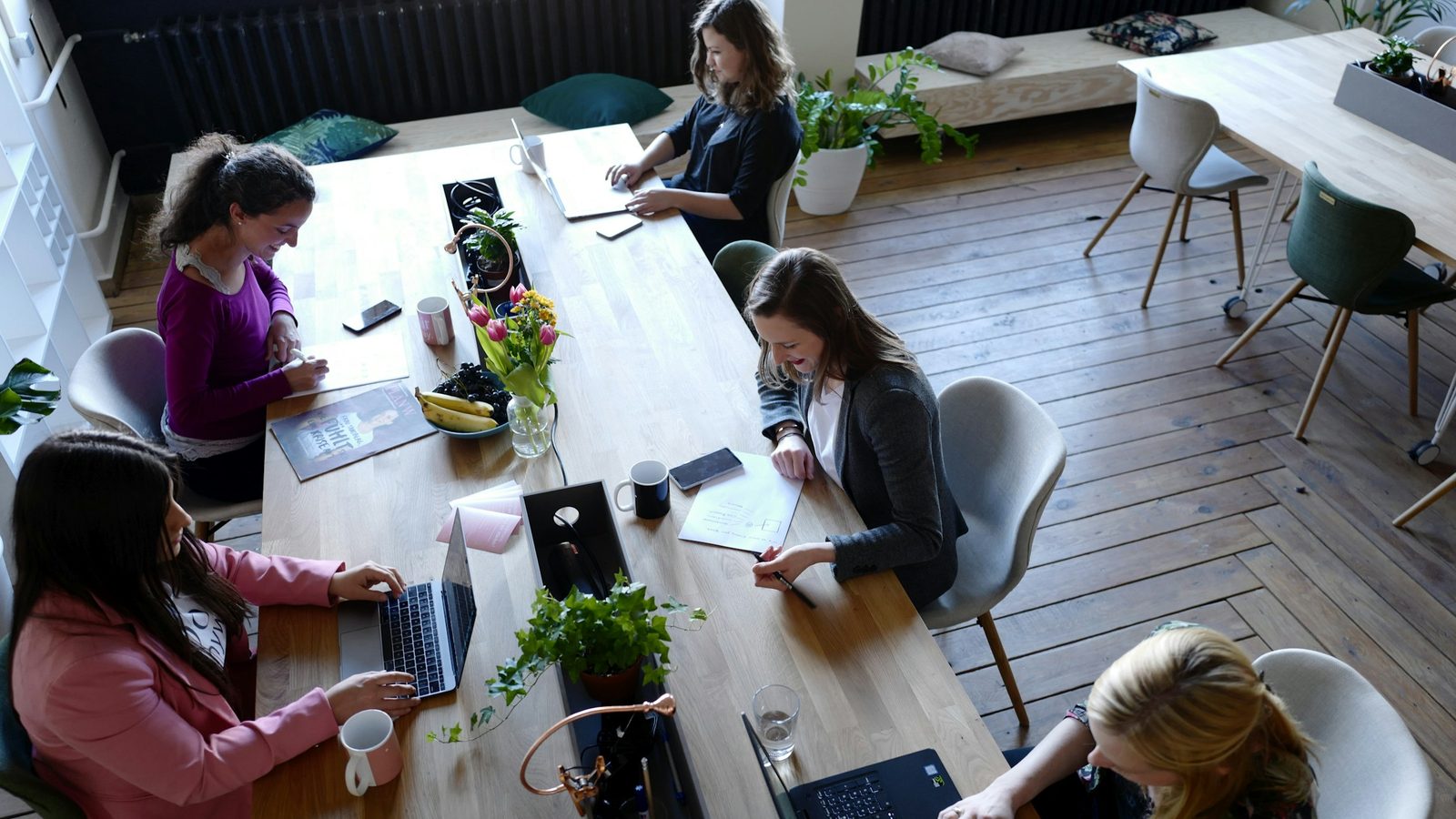 Women working on laptops and notebooks around a long wooden table in a bright shared office