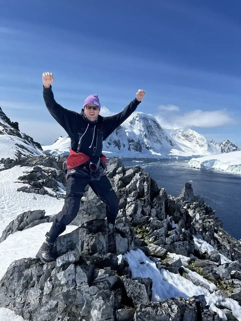 An expedition traveler celebrates on a snow-covered rocky ridge overlooking the Antarctic coastline. Surrounded by dramatic glaciers rugged peaks and deep-blue polar waters the hiker raises both arms in triumph after reaching a scenic viewpoint during an