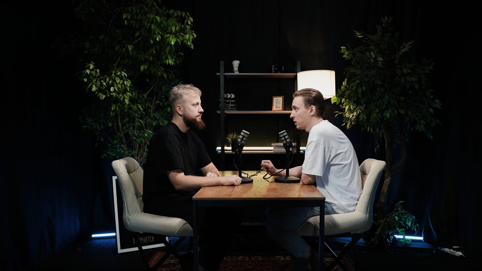 Двое мужчин записывают подкаст в студии с микрофонами RØDE PodMic, сидя друг напротив друга за столом, на фоне книжной полки, лампы и растений