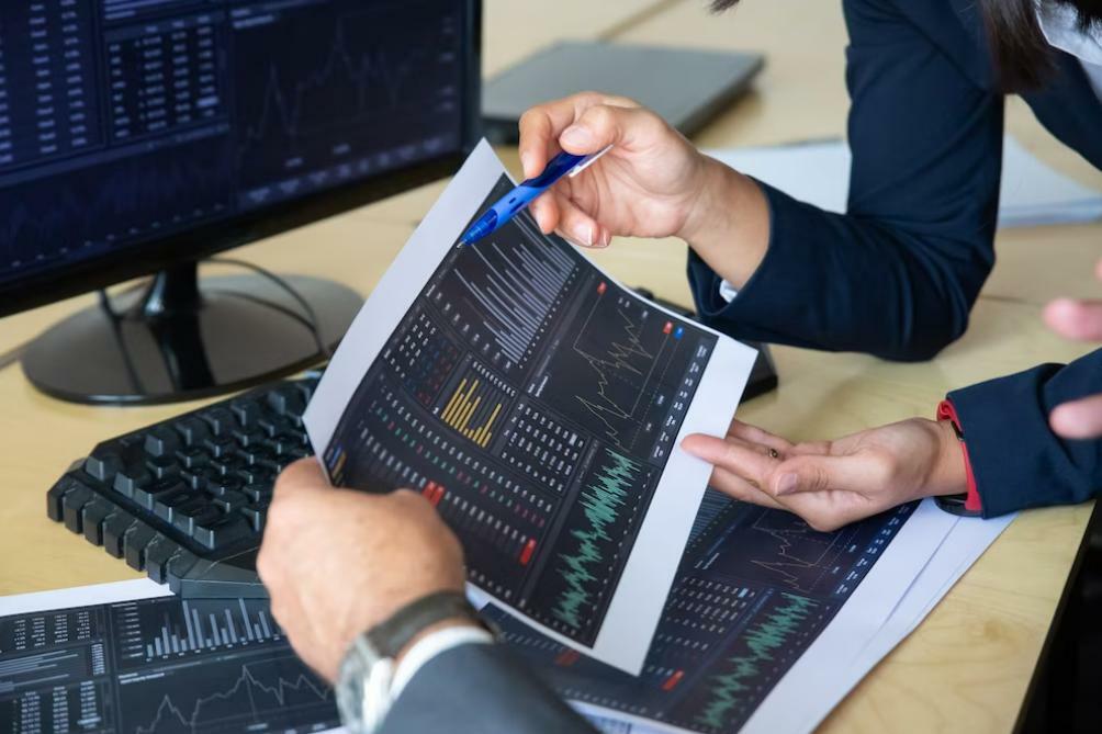 Vista parcial de los brazos de dos personas sosteniendo y analizando una imagen impresa de gráficos de precios para aprender trading