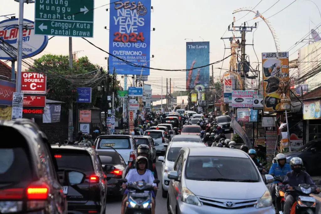 A candid daytime photo showing a heavily congested narrow street in Canggu. The road is completely packed with cars and dozens of motorbikes weaving through, illustrating the intense traffic jams often discussed by locals and tourists.