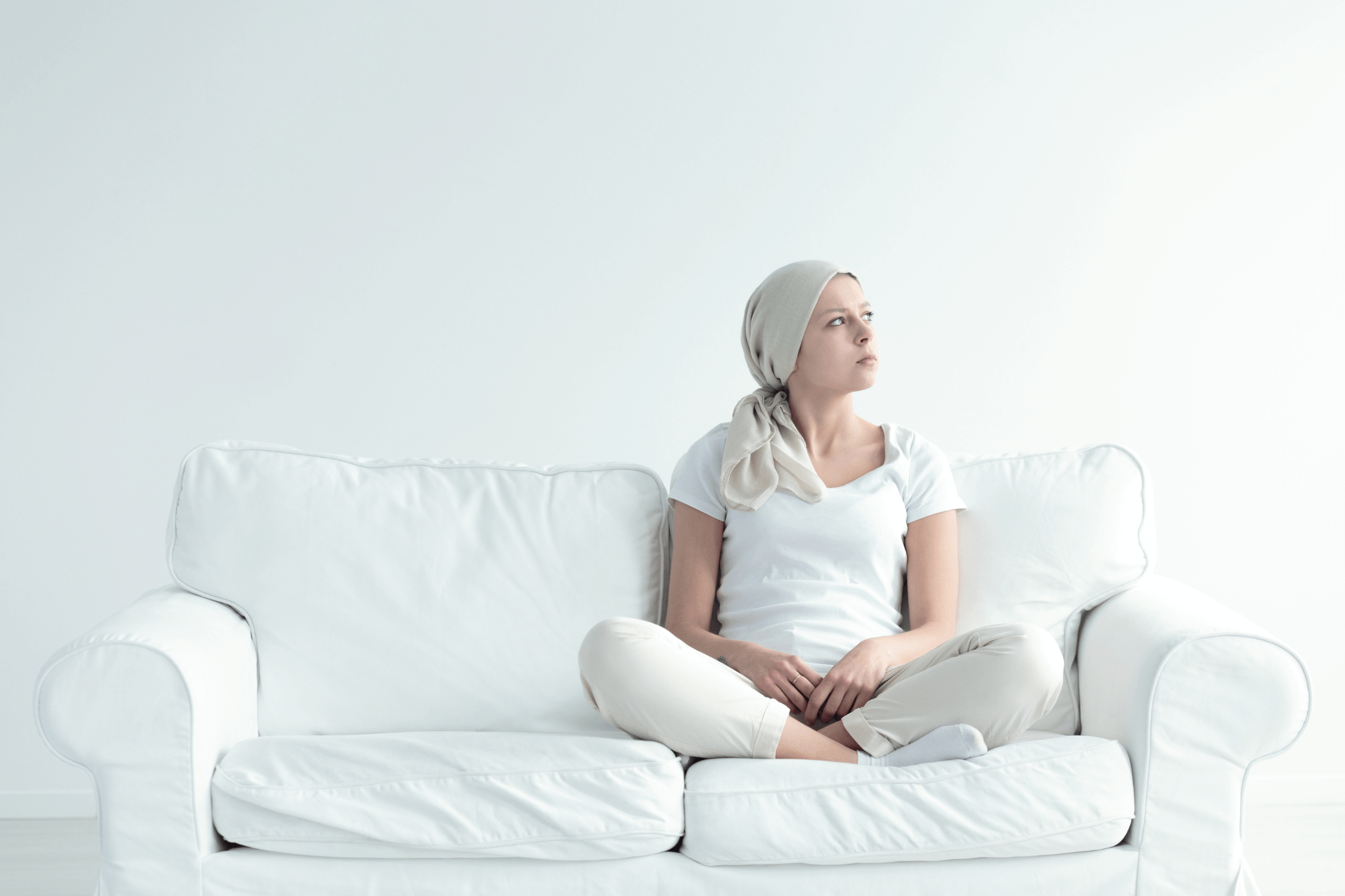 Nachdenkende Tumorpatientin mit Kopftuch sitzt weißbekleidet auf weißem Sofa