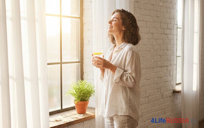 Женщина у окна в мягком утреннем свете держит стакан воды и делает глубокий вдох