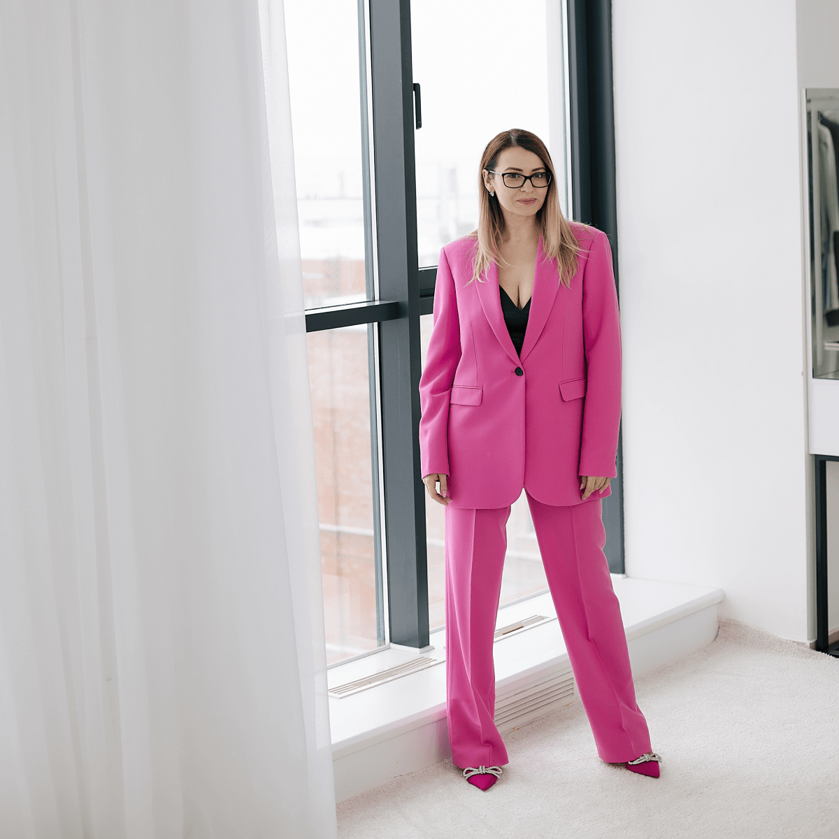 Девушка в очках стоит у окна в розовом костюме