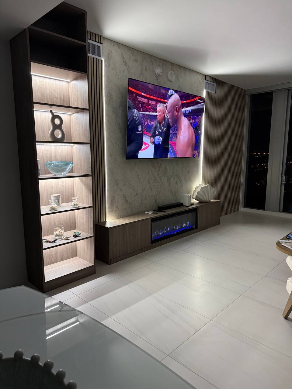Contemporary custom media wall with floating cabinetry, illuminated display shelves, decorative plaster wall finish, vertical wood slat accents, integrated LED lighting, and a linear electric fireplace in a luxury Miami residence.
