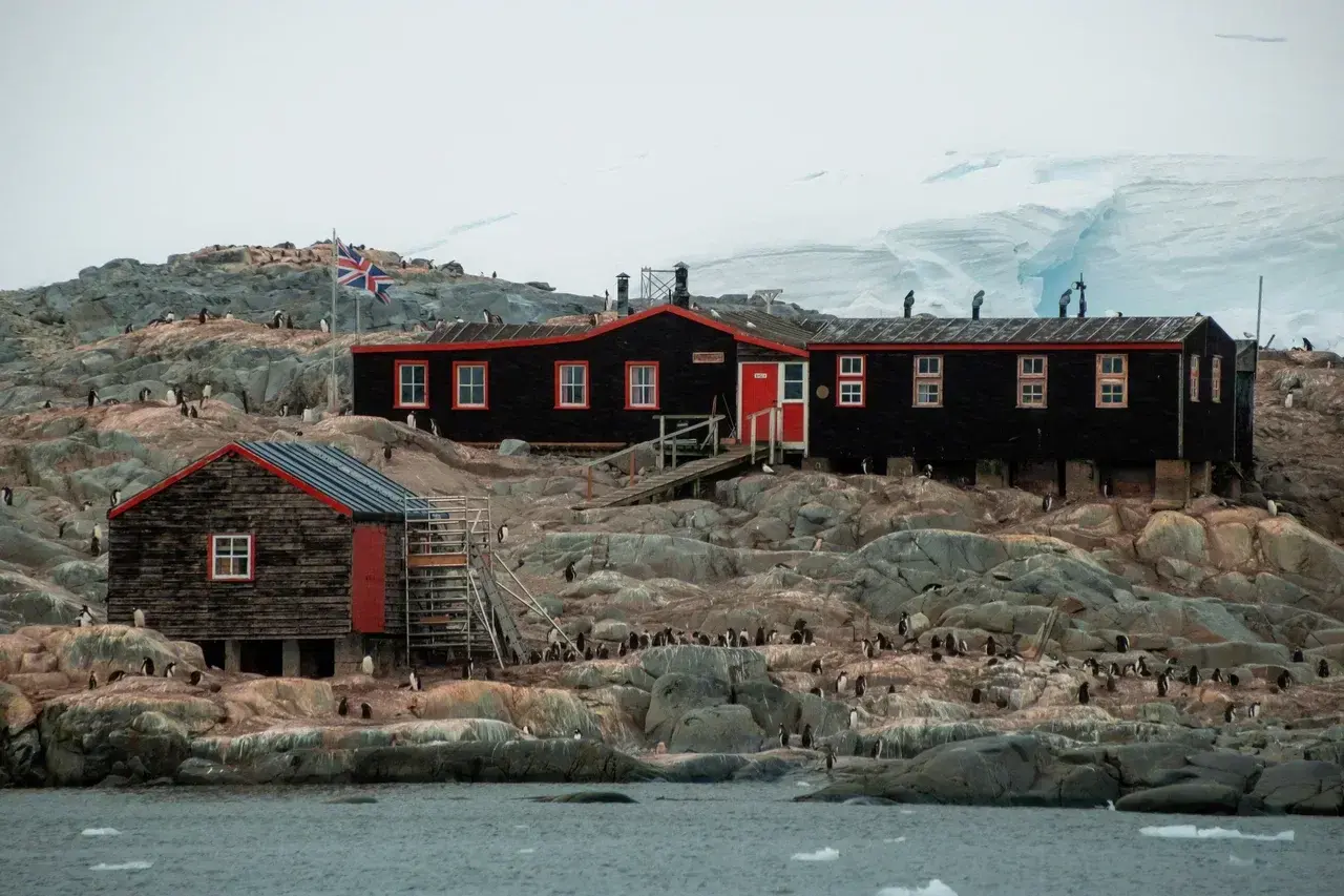 Historic Port Lockroy on the Antarctic Peninsula, a restored British research station and museum surrounded by hundreds of Gentoo penguins. The iconic black wooden buildings with bright red trim stand on a rocky shoreline beneath towering glaciers, offering visitors a glimpse into Antarctica's rich history of exploration while remaining one of the continent's most famous wildlife destinations.