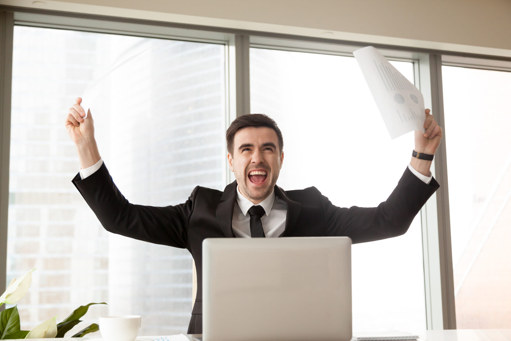 Overjoyed man in a business suit sitting at a desk with a laptop, triumphantly raising a document overhead, celebrating his new job offer.