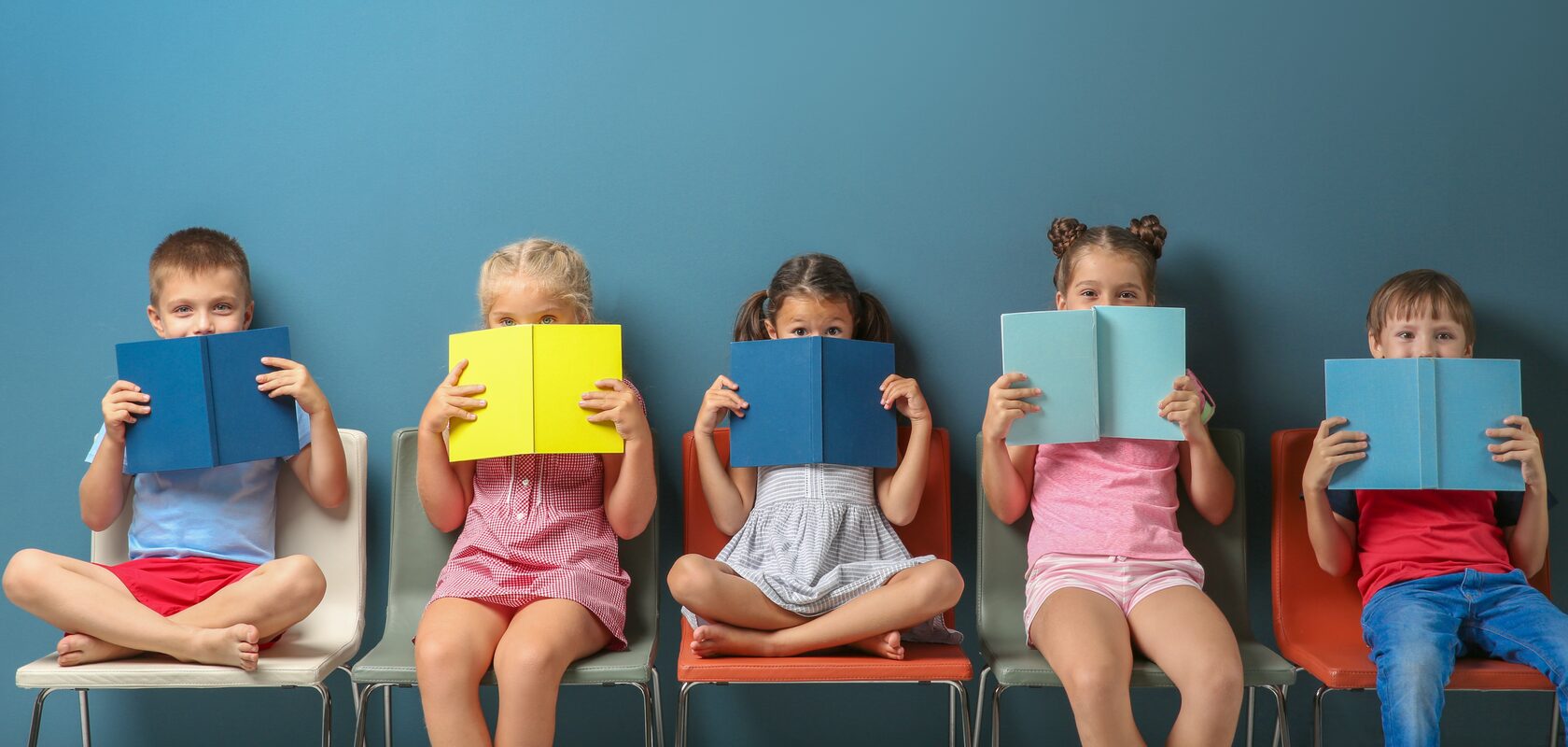 Children sitting in a line with their backs against a blue wall reading with their eyes peering over their books