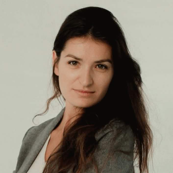 Портретная фотография молодой женщины с длинными темными волнистыми волосами, с лёгкой улыбкой, в сером пиджаке, надетом поверх белого топа, смотрящей прямо в камеру на фоне однотонной светло-серой стены.