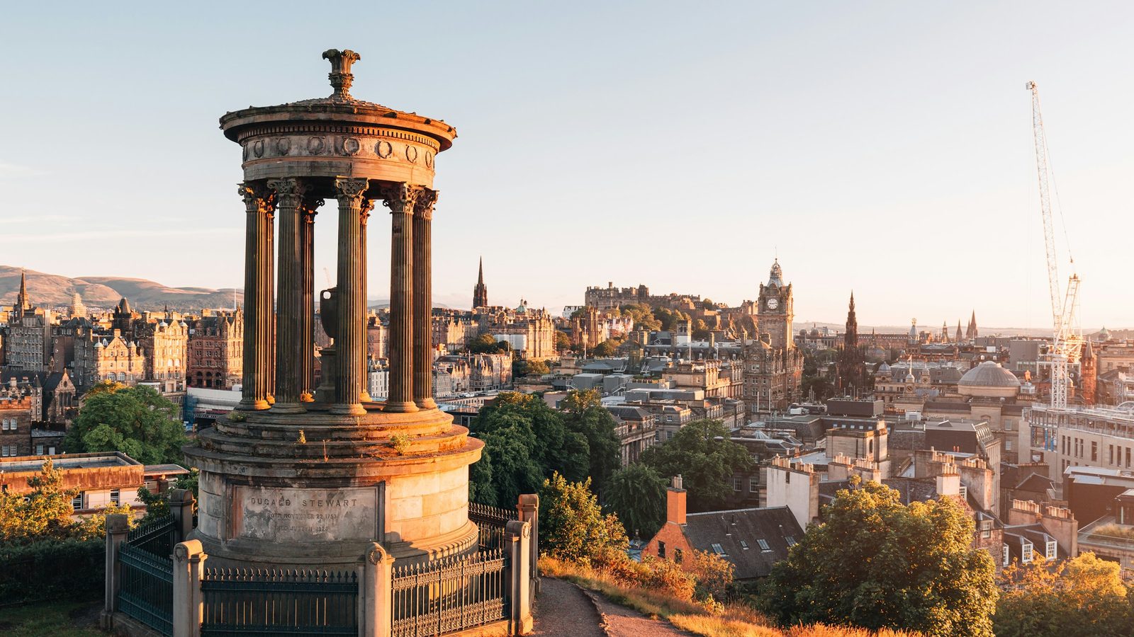 Edinburgh from Calton Hill at golden hour, with the Dugald Stewart Monument in front and Edinburgh Castle and the Balmoral clock tower behind