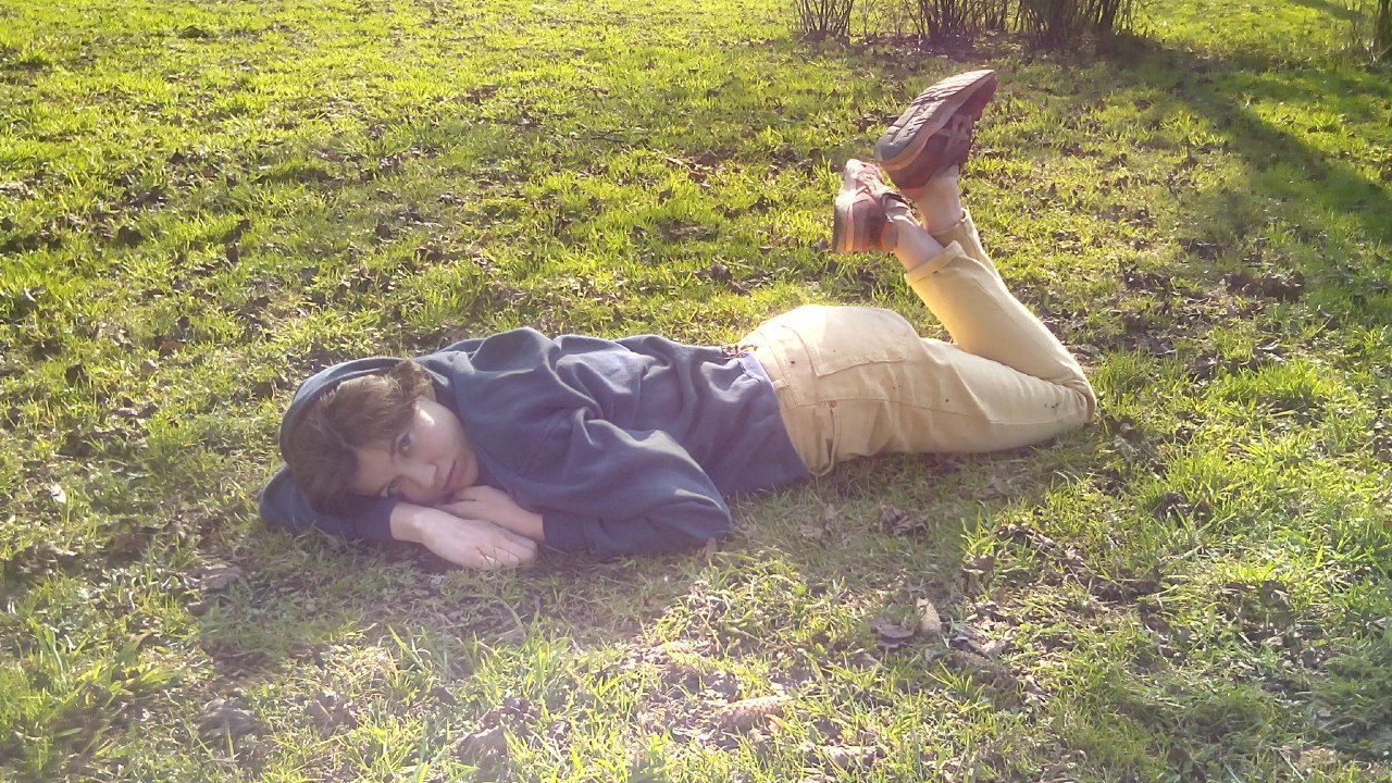 Тифлокомментарий. Цветная фотография. В солнечный день на траве лежит девушка и смотрит в камеру снизу вверх. Это Анна Терешкина. Она лежит на животе, положив голову на бок и сложив руки под головой. Анна согнула ноги в коленях и будто болтает ими в воздухе. Она одета в темно-серую толстовку с капюшоном, желтые узкие джинсы и бордовые кроссовки.